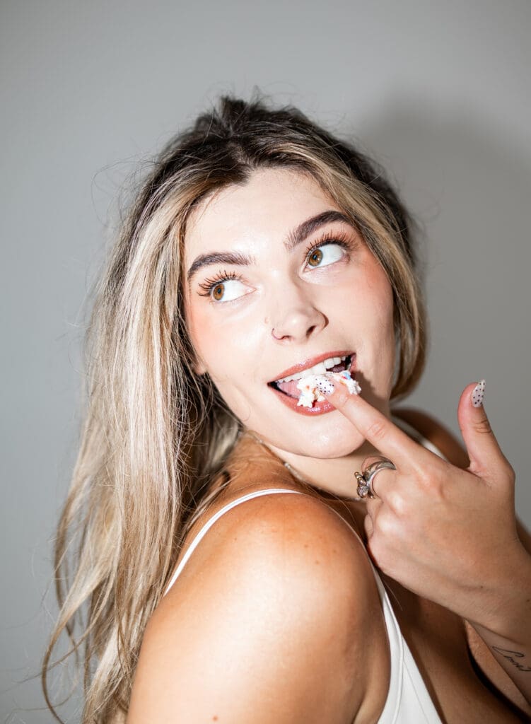 Woman with blonde hair and brown eyes licking birthday cake frosting off of her finger