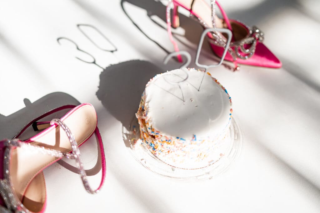 Pink statement heels with silver bow details thrown on the floor next to a white birthday cake with sprinkles and 30 candles