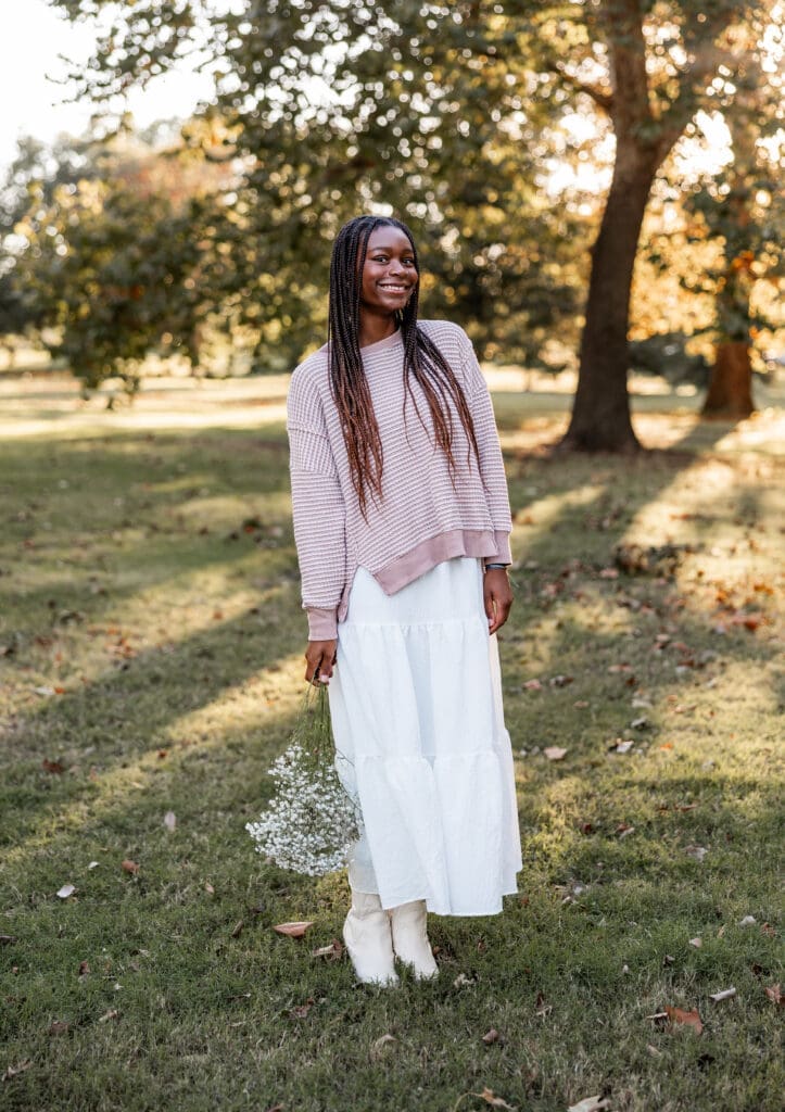 A young black woman with long hair wearing a white maxi skirt and striped sweater holds a bouquet of wildflowers during her Kansas City senior photoshoot