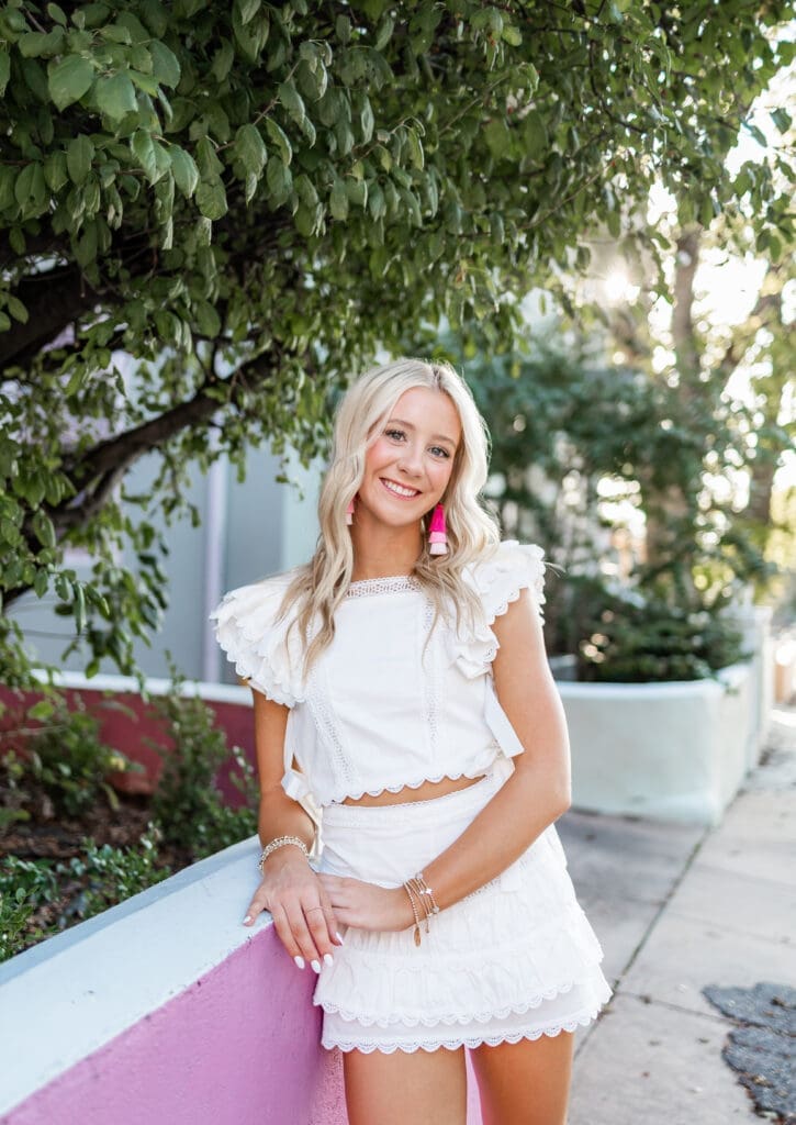 A woman with blonde hair leans against a pink wall while wearing a cute white skirt set with gold bracelets and pink statement earrings for her Overland Park senior photoshoot.