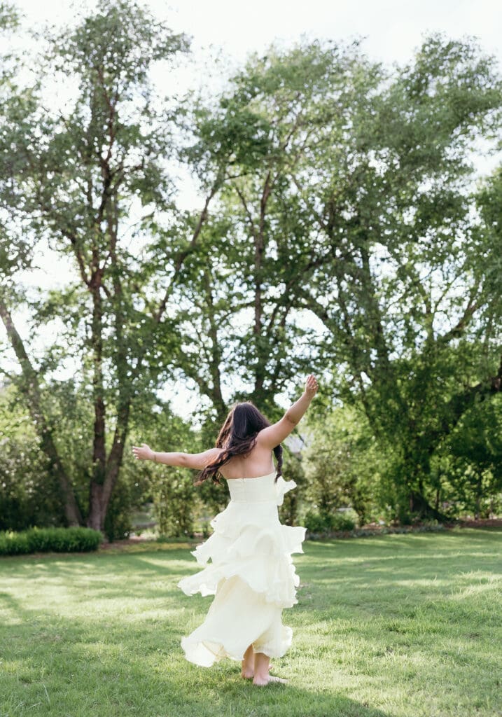 A young woman wearing a ruffled yellow maxi dress dances in a field of grass during her Kansas City senior photos.