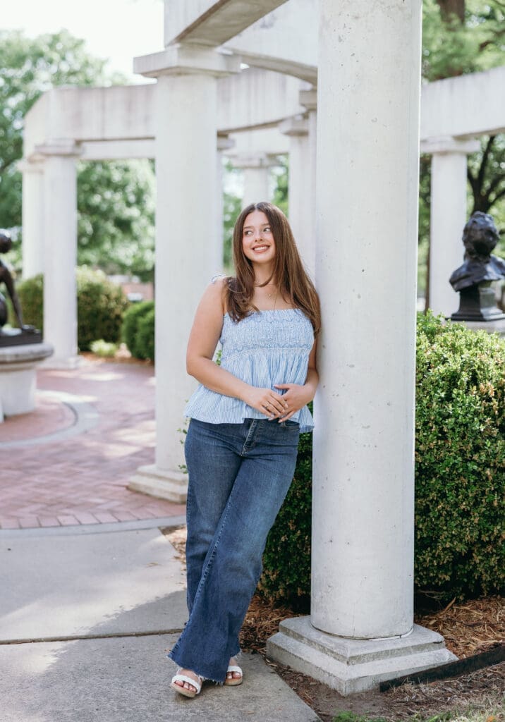 A young white woman with brown hair leans against a pillar while wearing blue jeans and a tank top during her Wichita senior pictures.