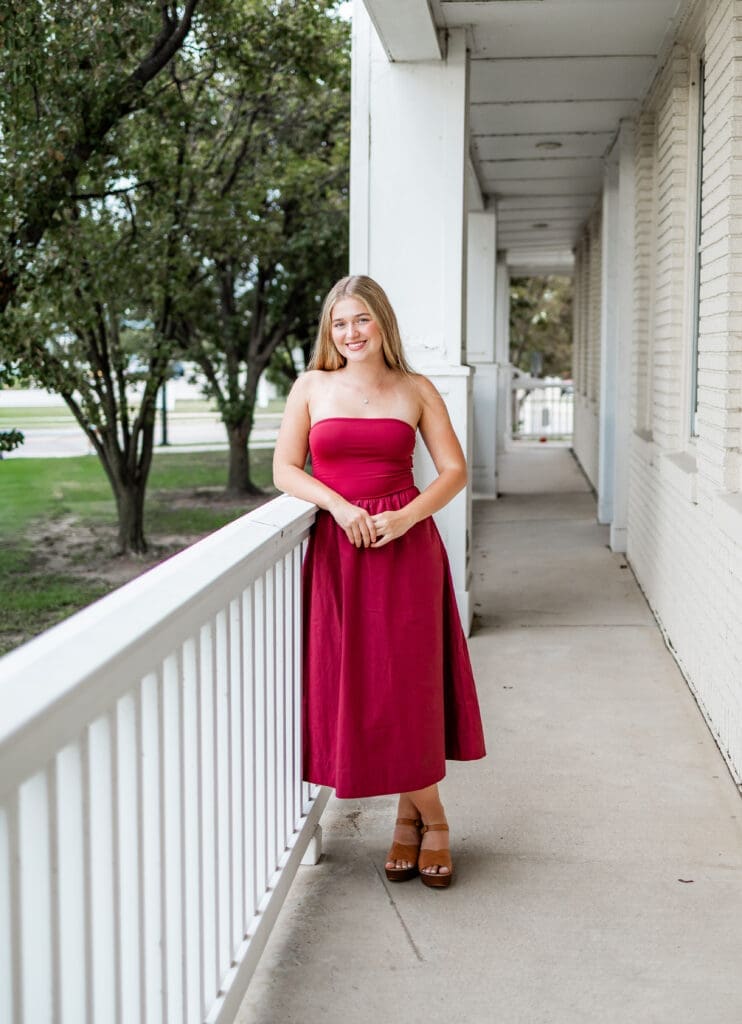 A blonde girl wearing a berry colored strapless dress leans on a white railing during her Wichita senior photos