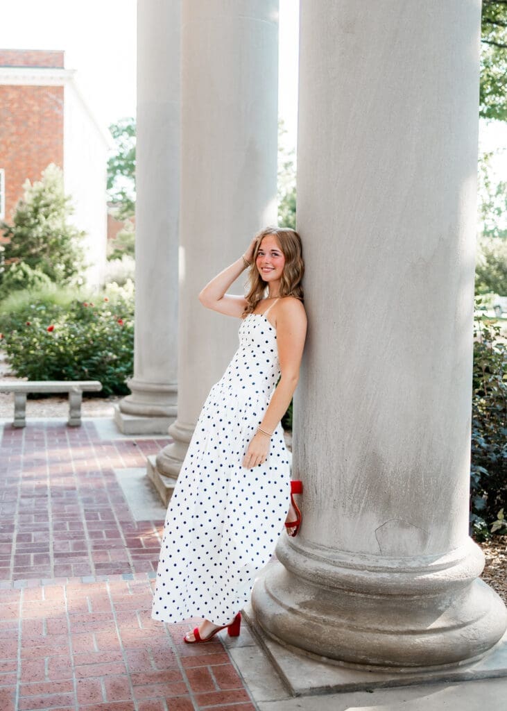 A girl wearing a black and white polk dot maxi dress and red heels leans against a pillar during her Kansas senior photos.