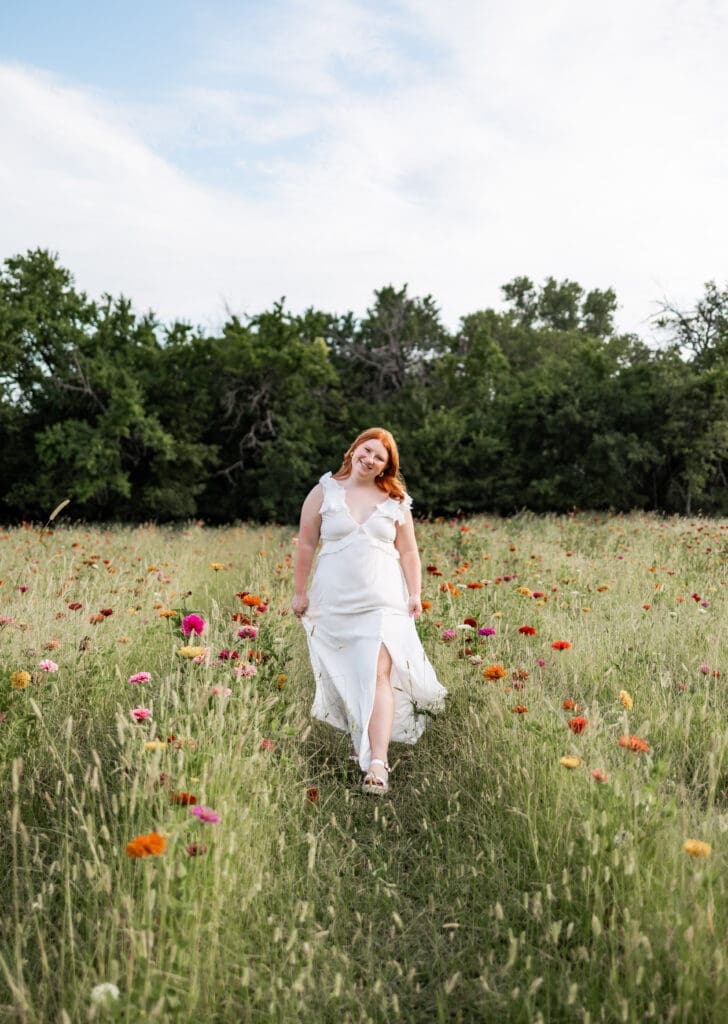 A girl with red hair wearing a long white dress walks through a field of wildflowers during her Olathe senior pictures