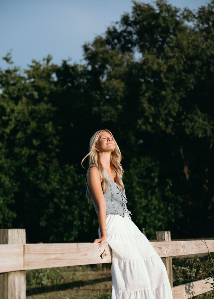 A woman with long blonde hair smiles at the sky while sitting on a wooden fence, wearing a long white skirt and a denim vest for her Kansas City senior pictures